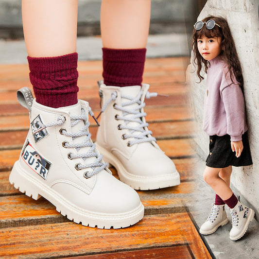 White boots with brand logo worn by a person on wooden floor, child in pink sweater and black skirt standing against a wall.