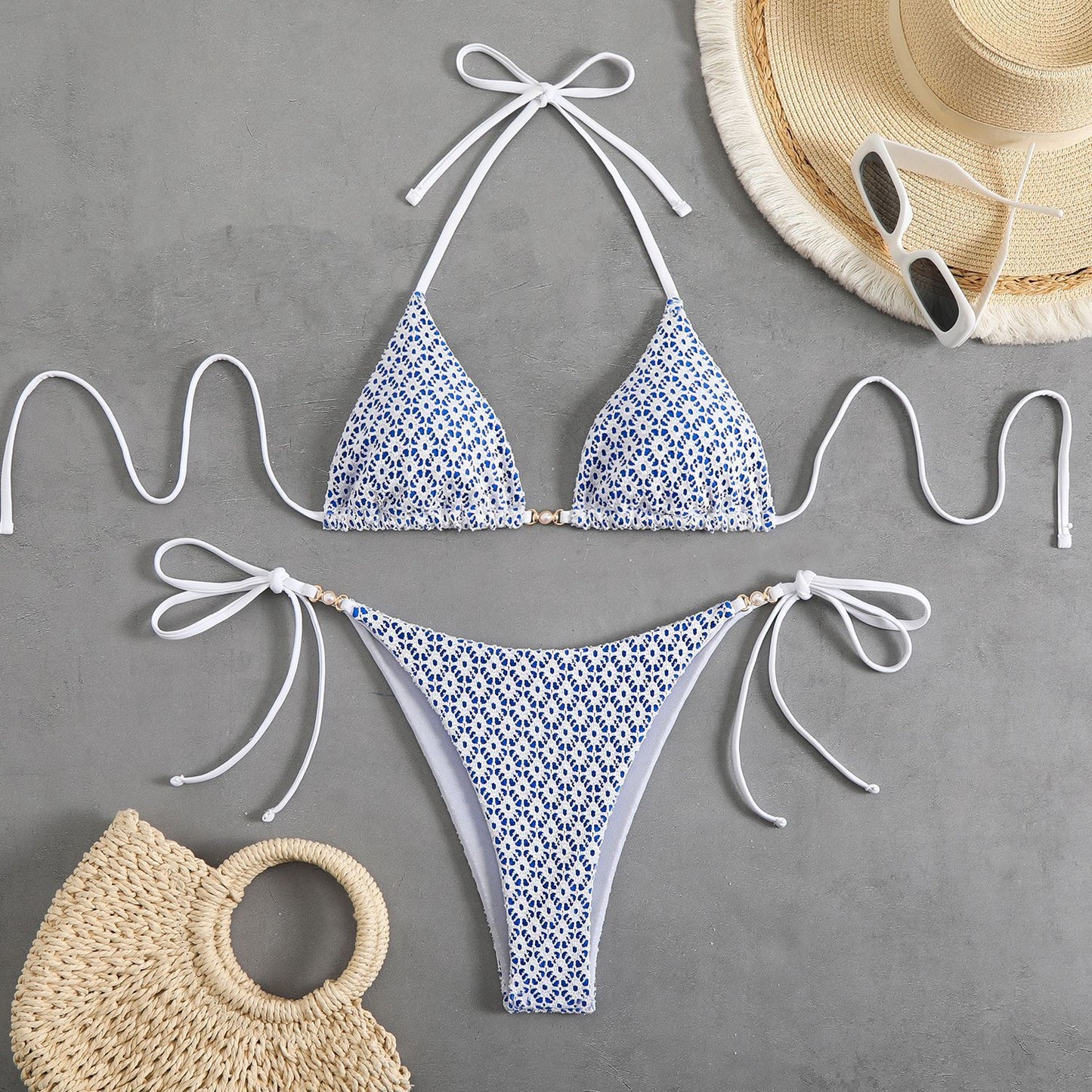 Blue floral bikini set with white straps on a gray background, accompanied by a straw bag, hat, and sunglasses.