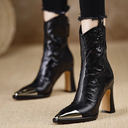 Black high-heeled boots with metallic toe detail on a beige surface