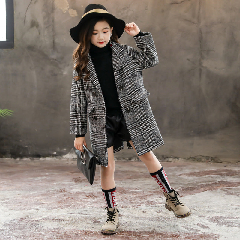 Young girl wearing a plaid coat and hat against a textured wall.