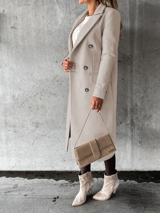 Woman wearing a long beige coat and white boots against a gray concrete wall.