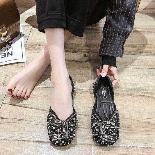Black embellished flats worn by a person on a wooden floor.