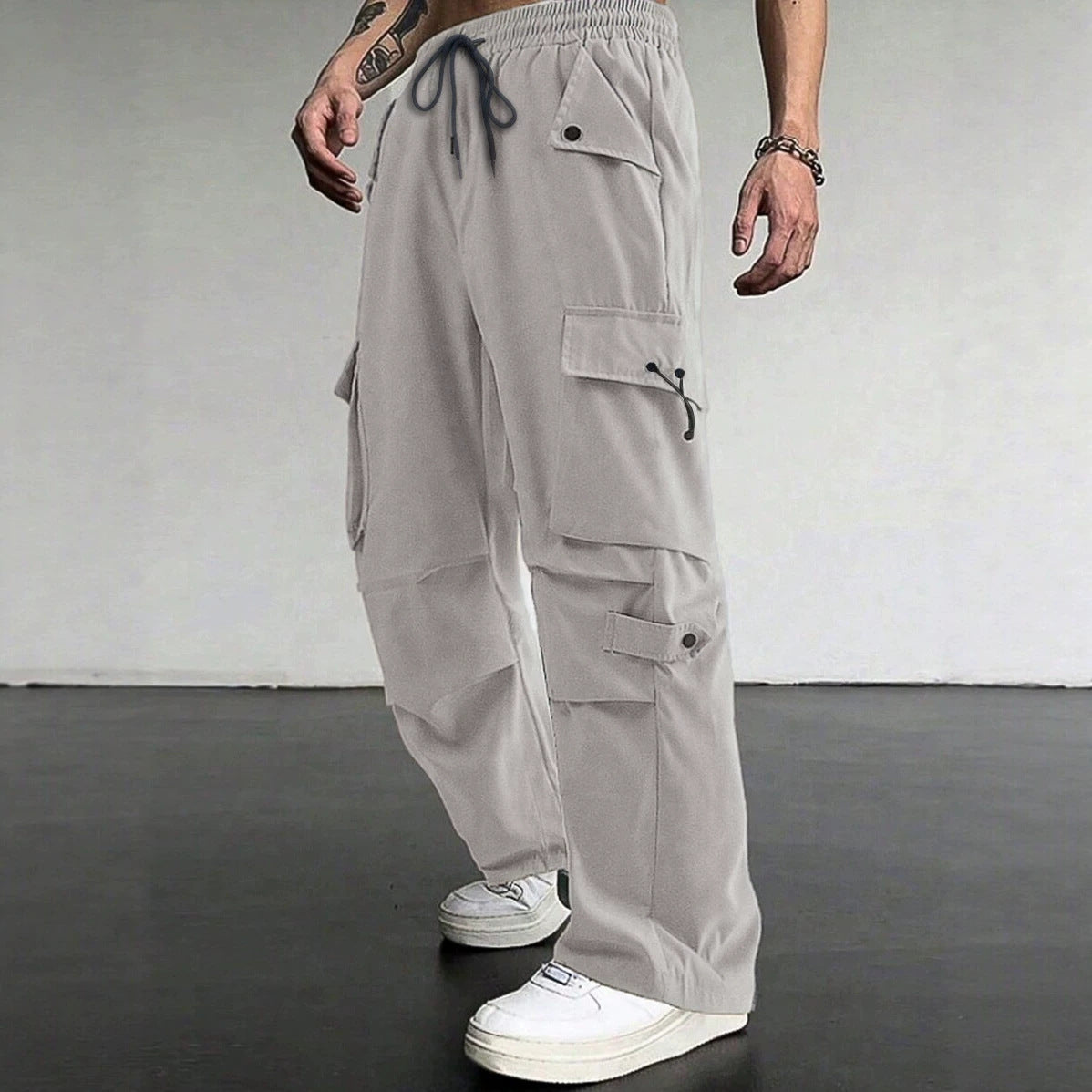 A person wearing light gray wide-leg cargo pants with multiple flap pockets, a drawstring waist, and white sneakers, standing against a plain light gray wall and dark floor.