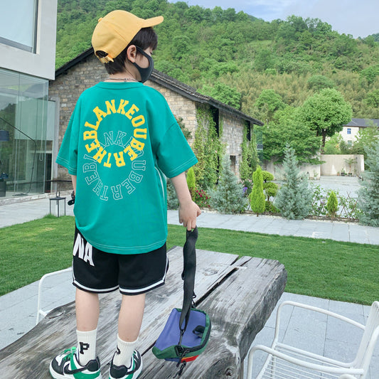 Child wearing a green t-shirt with text and black shorts, standing outdoors on a wooden platform.
