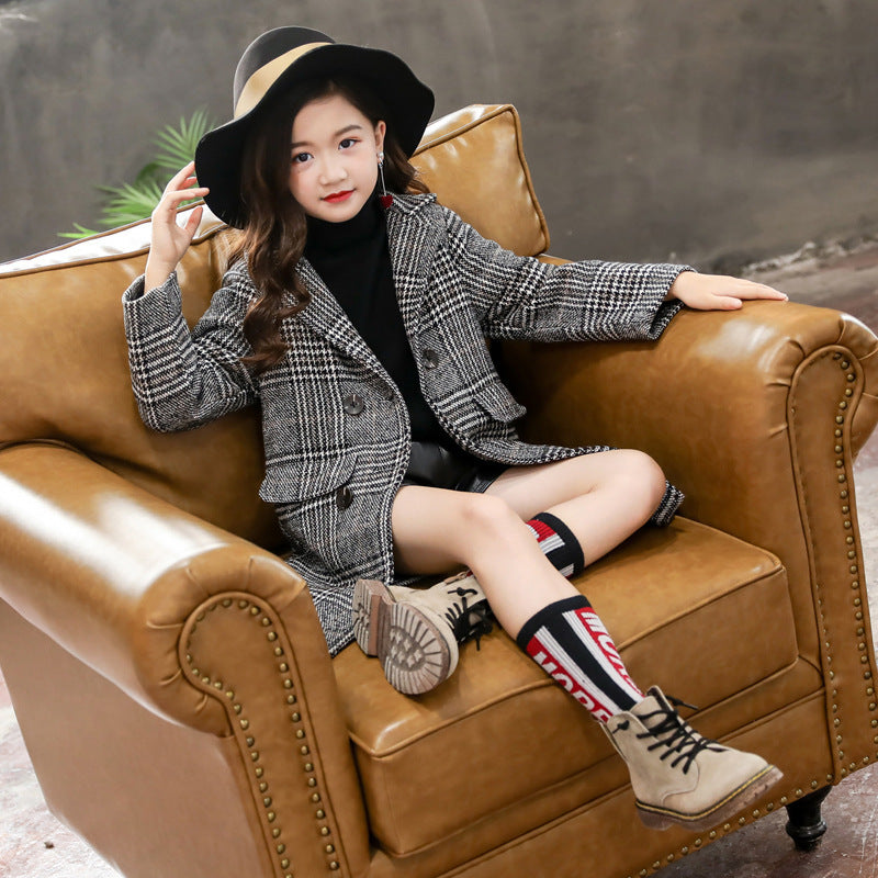Young girl sitting on a brown leather couch wearing a gray plaid jacket and black hat.