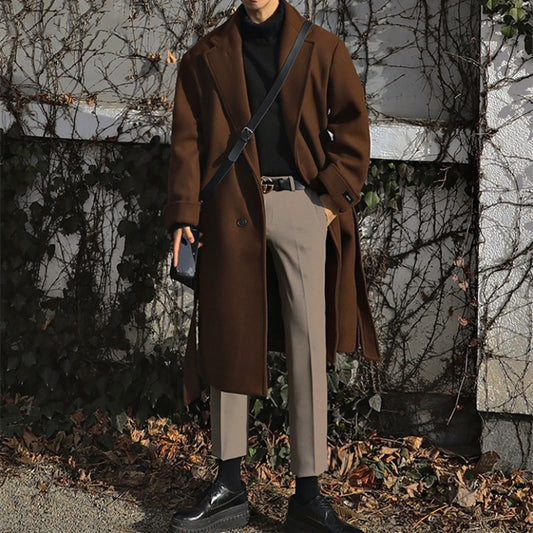 Person wearing a brown coat standing outdoors with a textured wall and plants in the background
