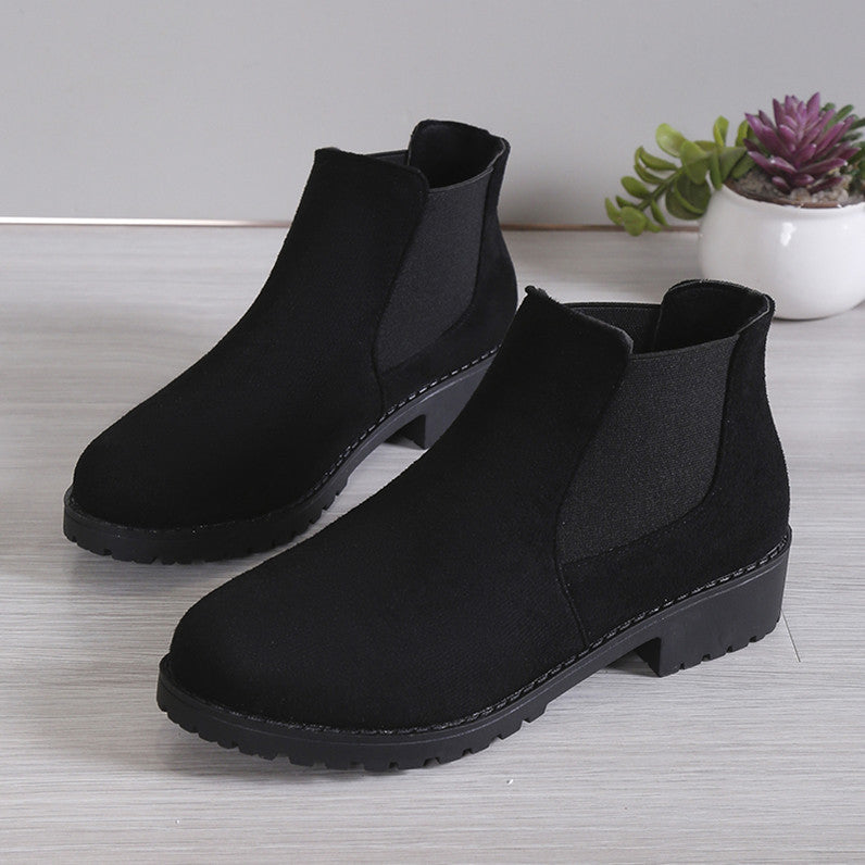 Pair of black ankle boots on a light wooden floor with a plant in the background.
