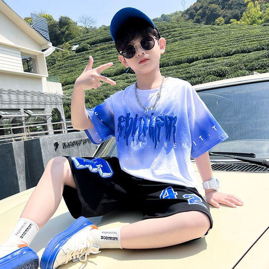Child wearing a blue and white t-shirt with text, black shorts with a logo, and blue sneakers, sitting on a car with a scenic background.
