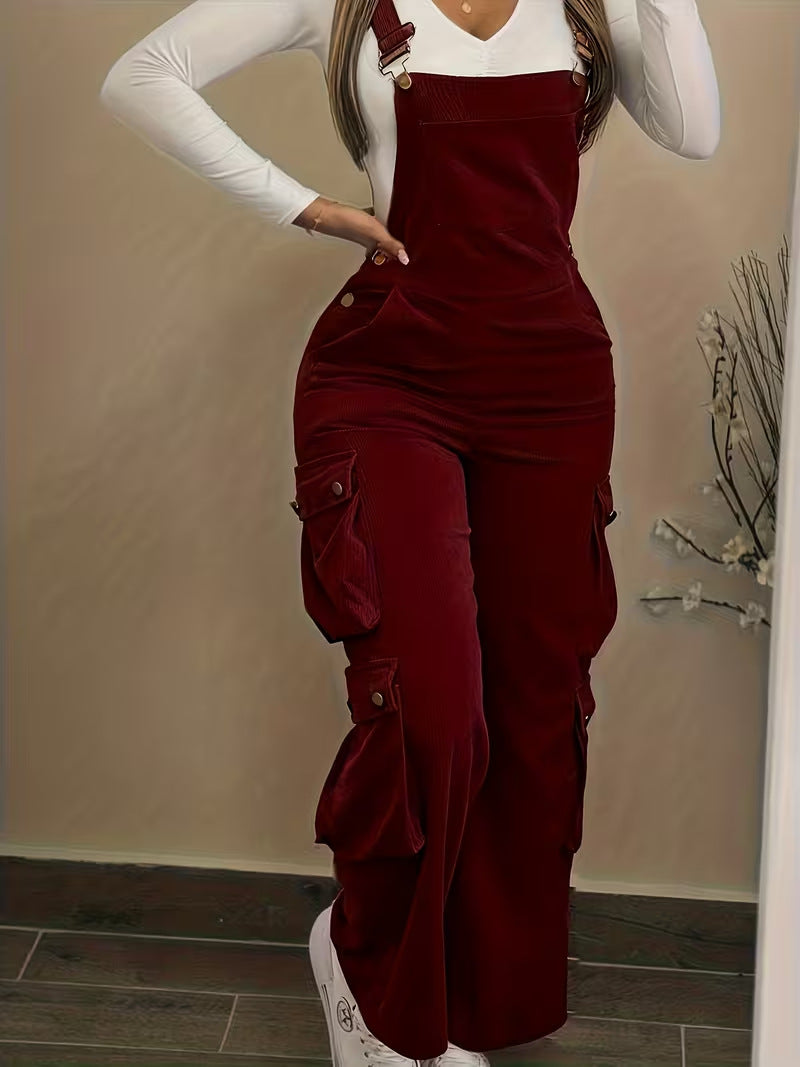 Person wearing a burgundy corduroy overalls with a white long-sleeve shirt underneath, standing against a beige wall.