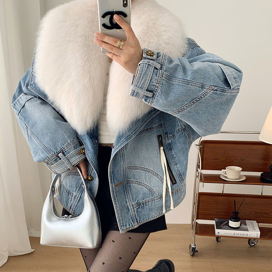 Person wearing a denim jacket with a white fur vest, holding a phone, in a room with a side table and lamp.
