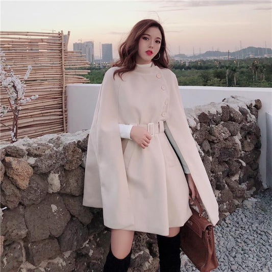 Woman in a beige coat standing against a stone wall with a scenic background