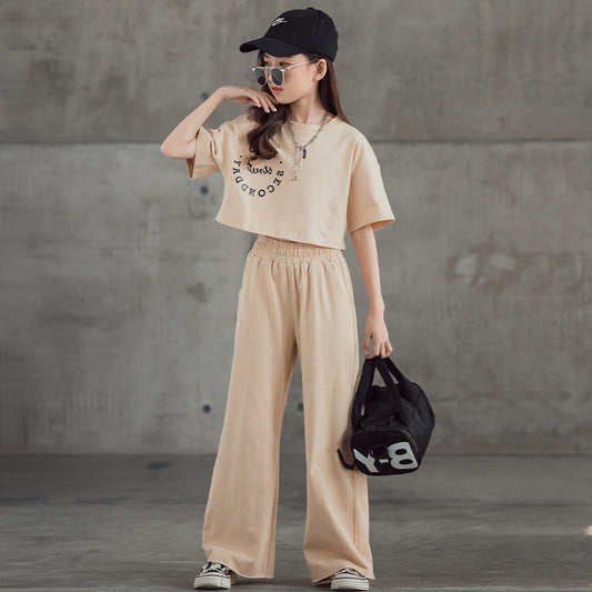 Person wearing a beige outfit with wide-leg pants and a cap, holding a black bag against a gray concrete wall.
