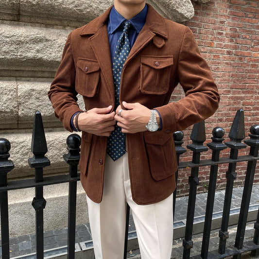 Man wearing a brown jacket, blue shirt, and white pants standing against a brick wall.