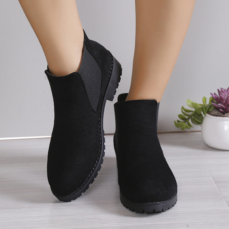 Black ankle boots worn by a person on a light wooden floor with a plant in the background