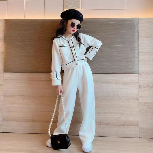 Person wearing a white outfit with black accents, holding a black handbag with a pearl strap, against a wooden wall.