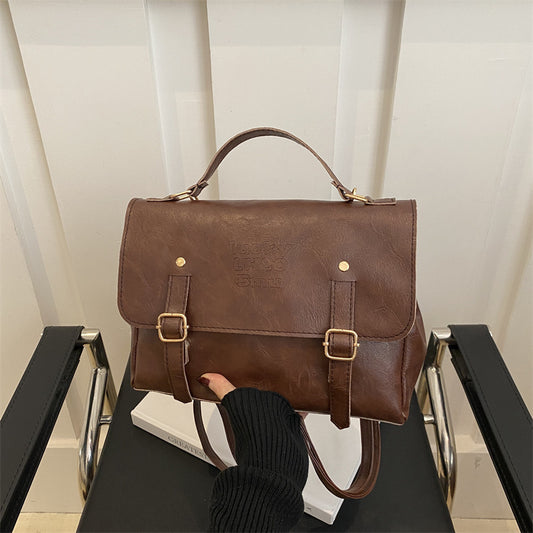 Brown leather satchel bag held by a person on a black chair with a white curtain background