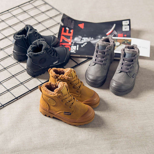 Three pairs of children's boots in yellow, black, and gray on a textured surface with a magazine underneath.