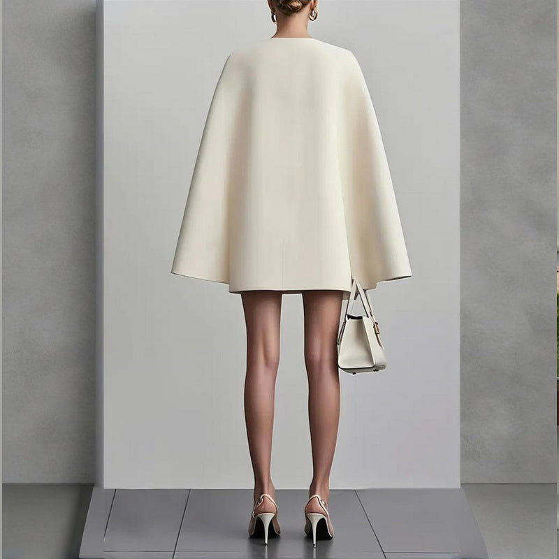 Person wearing a cream-colored cape over a dress, holding a matching handbag against a plain background.