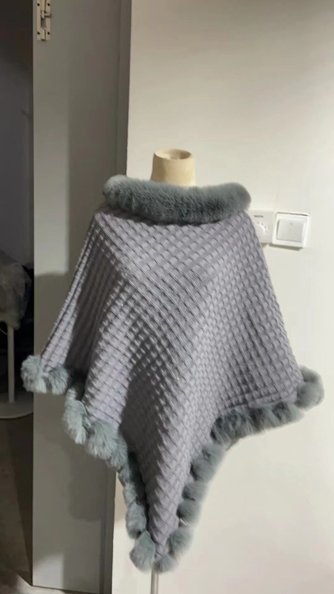 Gray knitted poncho with pom-poms on a mannequin against a neutral wall.