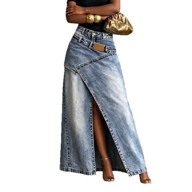 A model wearing a high-waisted, light-wash blue denim maxi skirt with a diagonal front slit and a brown leather patch detail. The model is also wearing a black top, gold bracelets, and holding a metallic gold clutch against a plain white background.