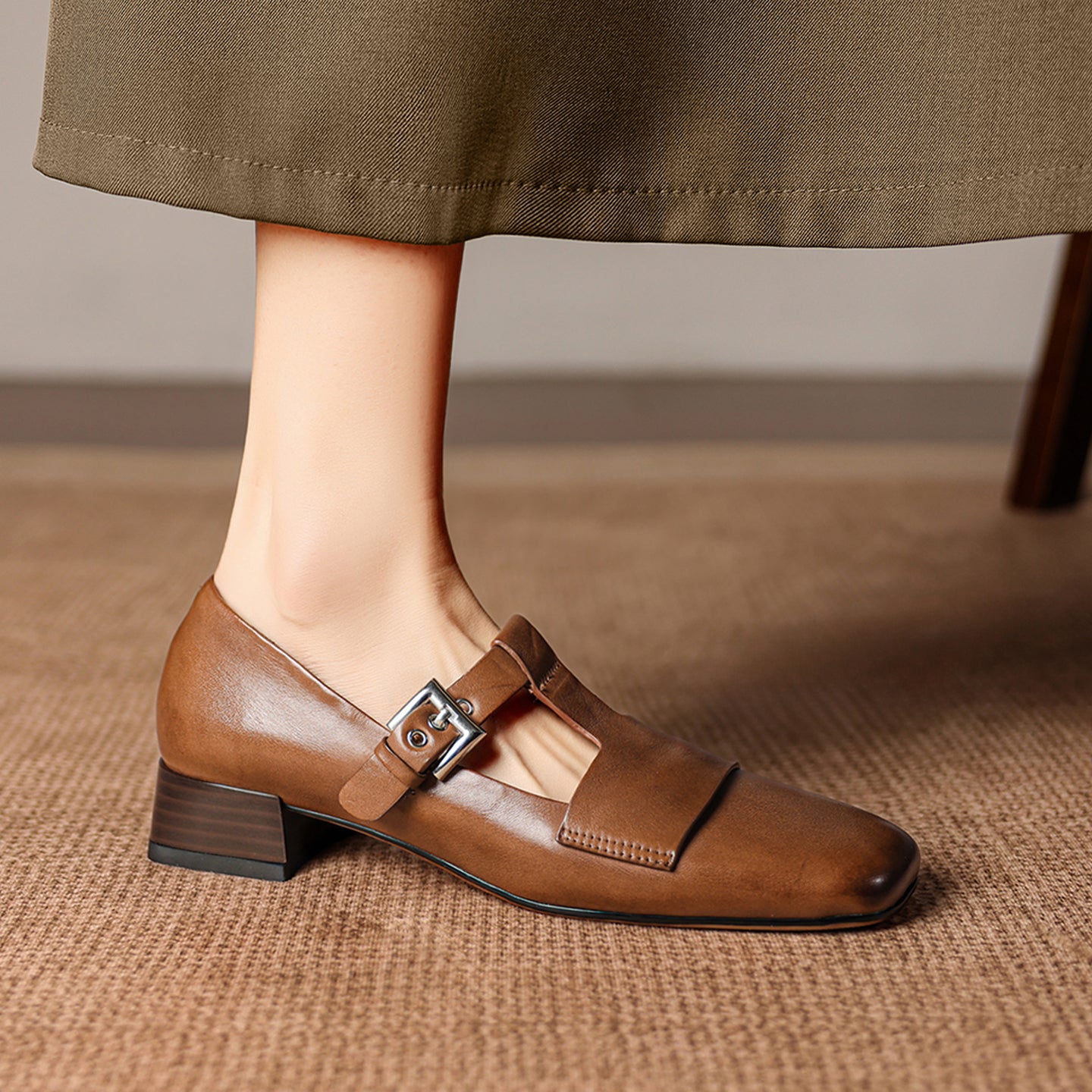 Brown leather shoe with a buckle worn by a person on a textured surface.