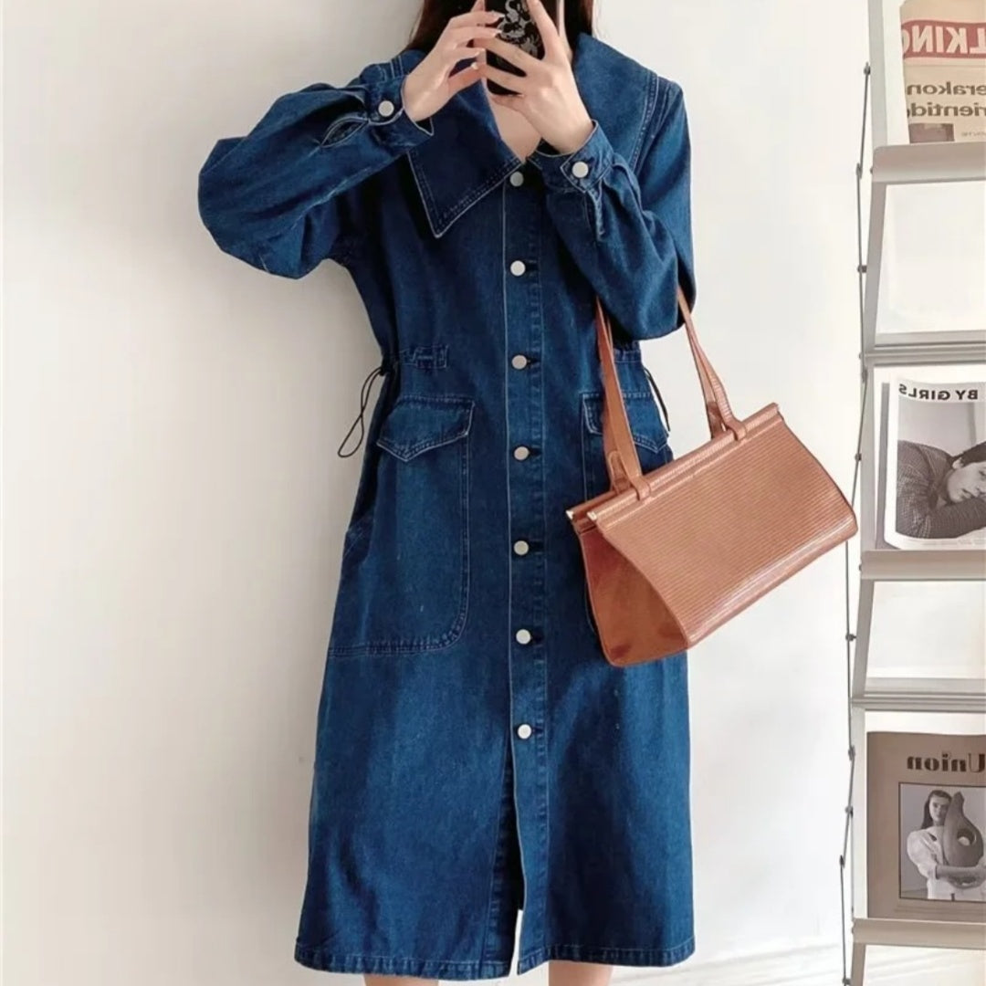 Person wearing a blue denim dress holding a brown handbag against a white wall.