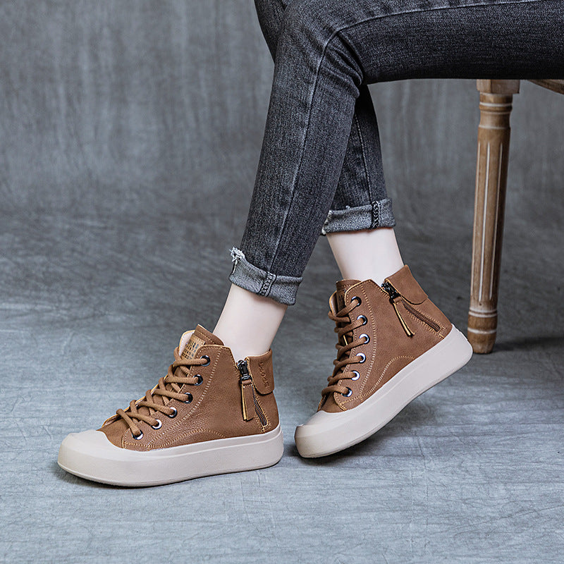 A person seated on a wooden chair wearing brown suede high-top sneakers with off-white soles and laces, paired with dark gray cuffed jeans, against a gray studio backdrop.