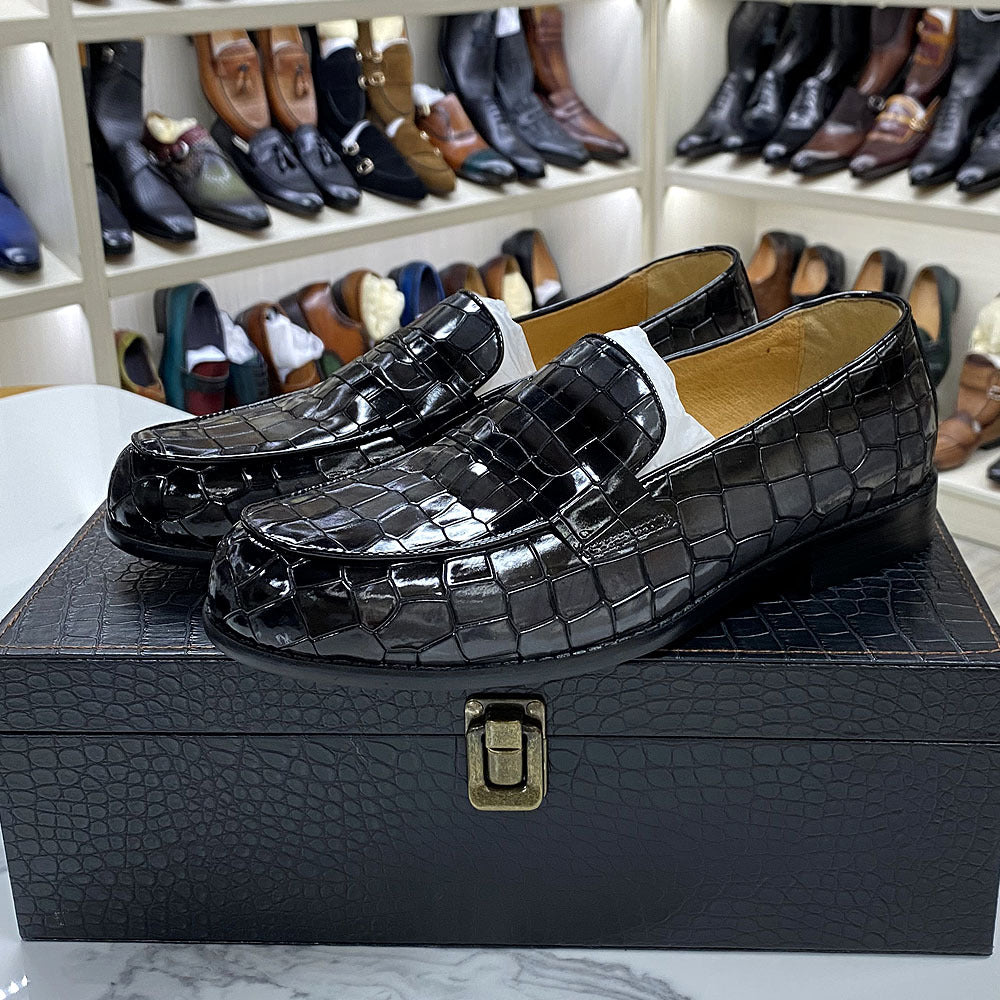 Black crocodile-patterned loafers on a black textured box with shoes displayed in the background.