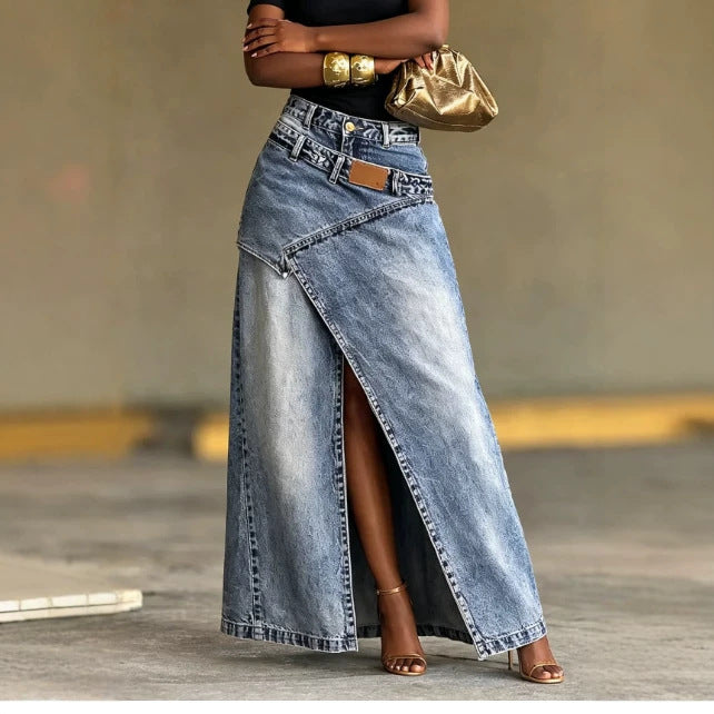 A woman models a high-waisted, light-wash blue denim maxi skirt with an asymmetrical wrap front and a thigh-high slit. She is wearing a black top, gold bracelets, a metallic gold clutch, and brown heeled sandals, standing against a neutral concrete background.