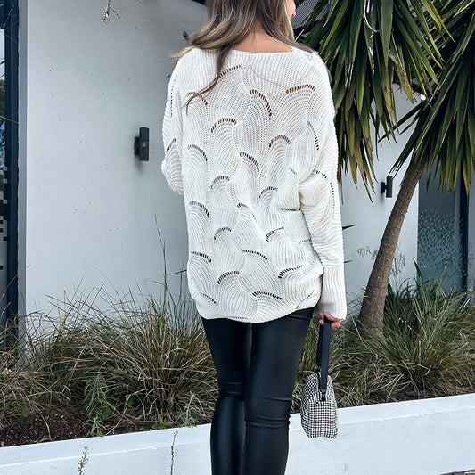 Person wearing a white patterned sweater and black pants, holding an umbrella, standing in front of a white wall with plants.