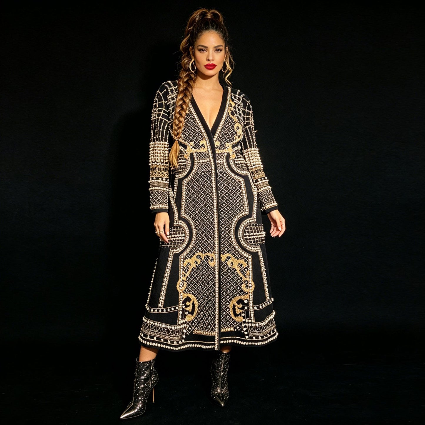 A model stands against a black background wearing a long black wrap-style dress heavily embellished with white and gold beadwork and patterns, paired with black pointed-toe ankle boots.