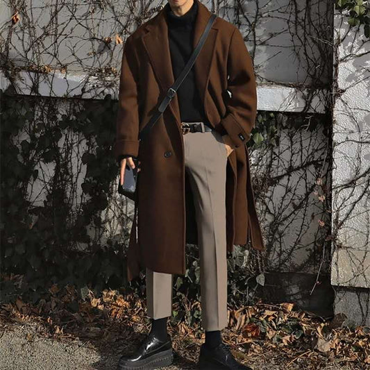 Person wearing a brown coat and holding a phone, standing against a backdrop of bare trees and a building.
