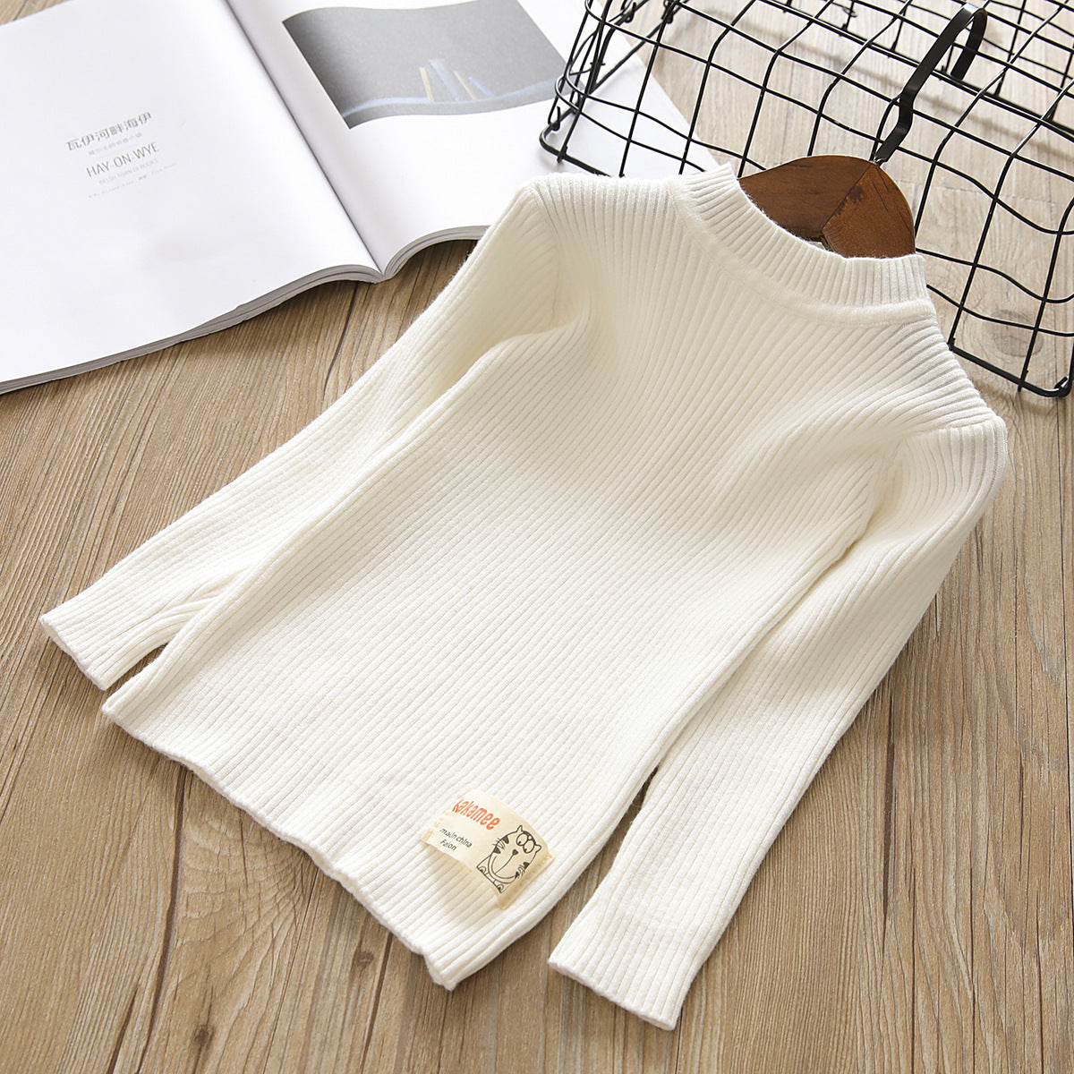 White ribbed sweater on a wooden surface with a magazine and black wire basket in the background.