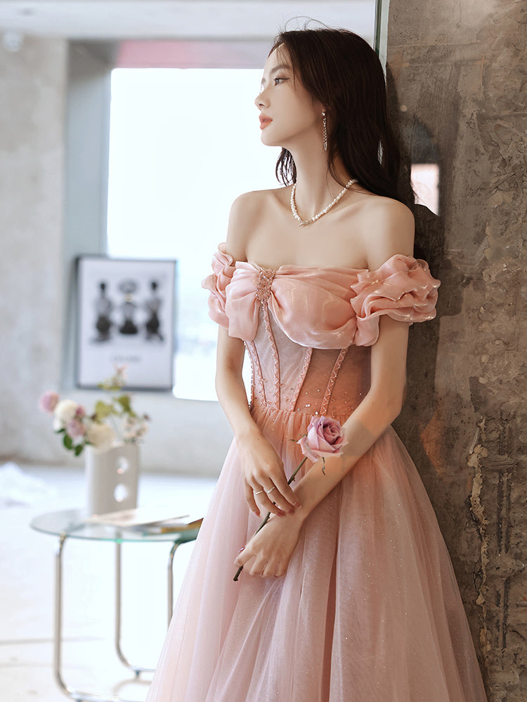 Woman in a pink dress holding a flower, standing against a stone wall with a blurred indoor background.