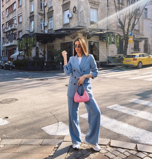 Woman in a light blue suit with a pink handbag crossing a street in an urban setting. by millivan