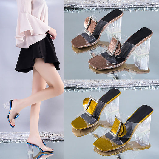 Transparent high-heeled shoes with gold and black accents worn by a person on a reflective surface.