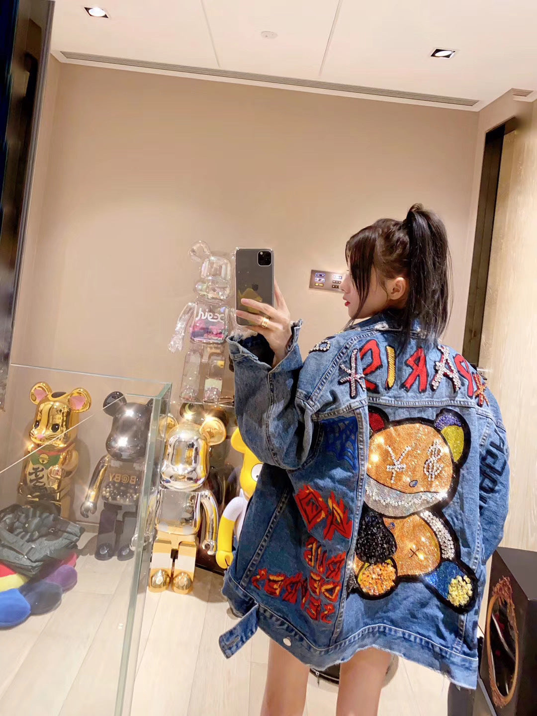 Person wearing a colorful denim jacket with cartoon designs, taking a mirror selfie.