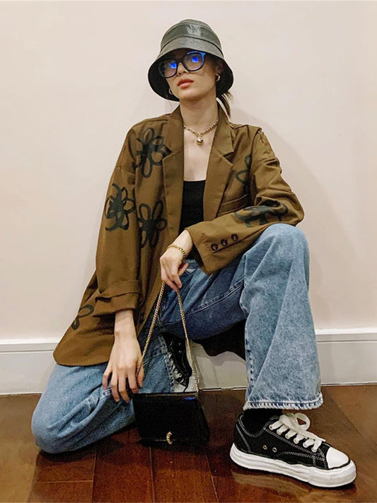 Person wearing a brown jacket with graphic designs, blue jeans, black sneakers, and a bucket hat. by millivan