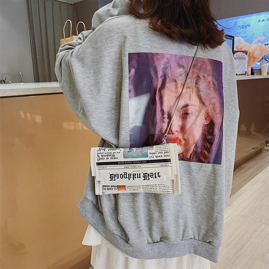 Person wearing a gray hoodie with a graphic design of a woman's face on the back, holding a newspaper.