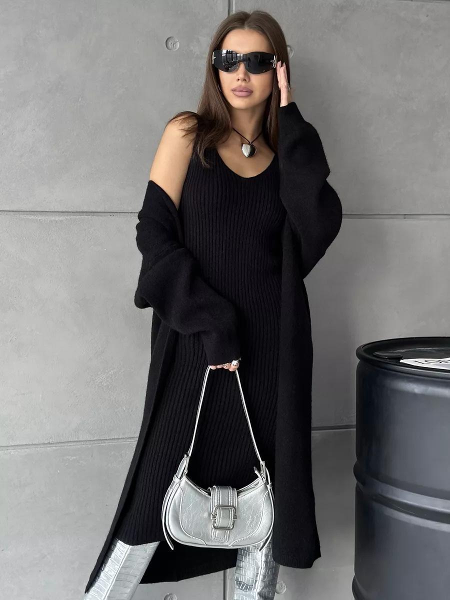 Woman in black outfit with sunglasses and a silver handbag against a gray wall.