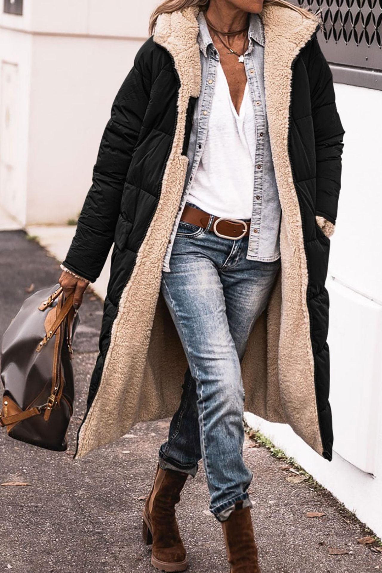 Person wearing a long black and beige coat with blue jeans and brown boots on a street.