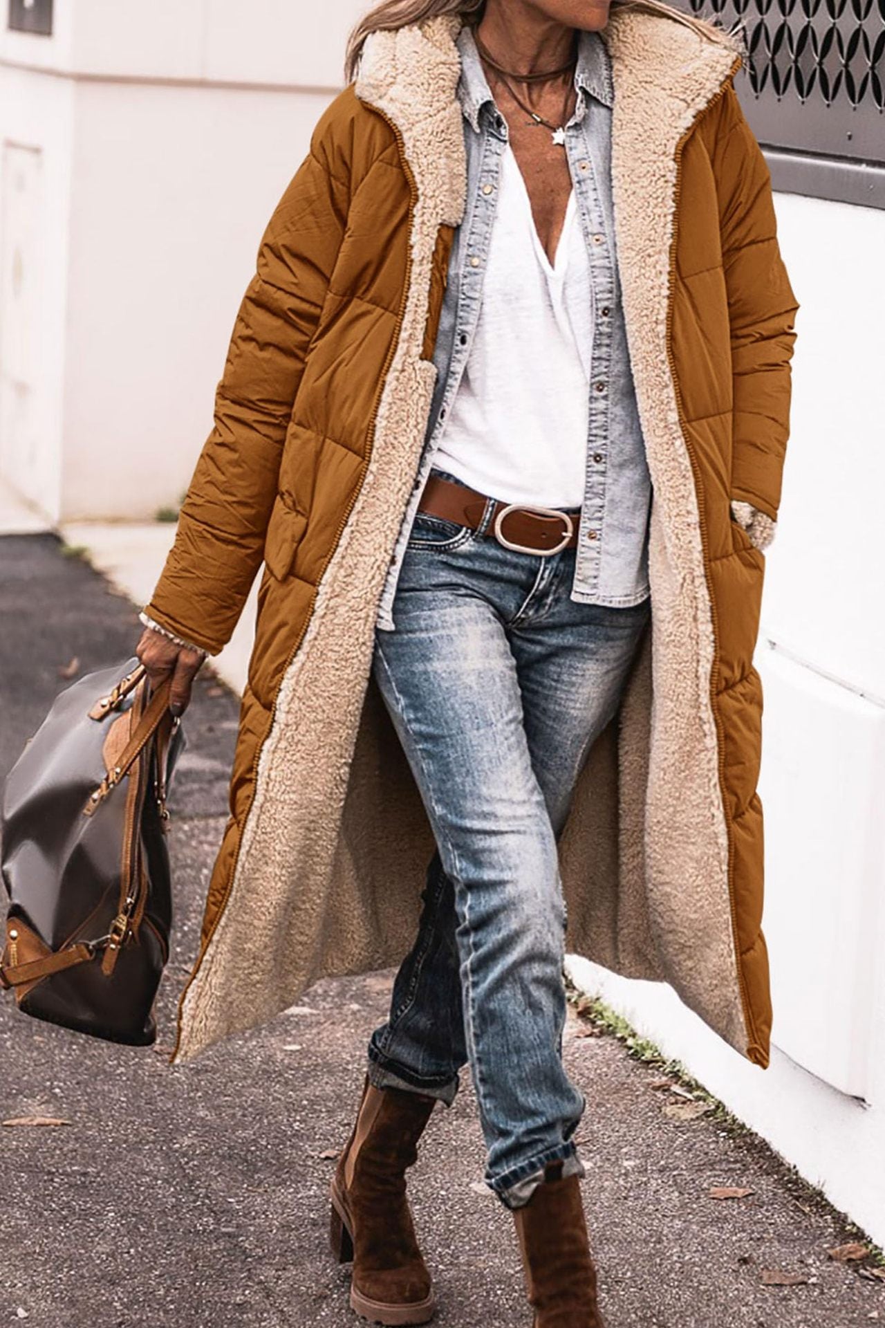 Person wearing a long brown puffer coat with a white shirt and blue jeans, standing on a street.