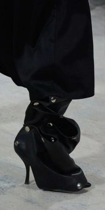 Black high-heeled shoes with studded details worn by a person in a black outfit on a light gray background.