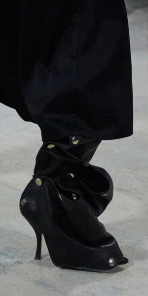 Black high-heeled shoes with studded details worn by a person in a black outfit on a light gray background.