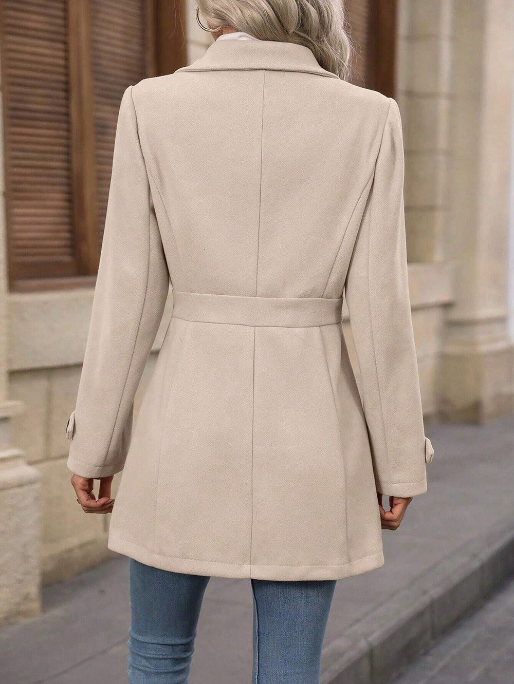 Person wearing a beige coat standing on a street with a building in the background
