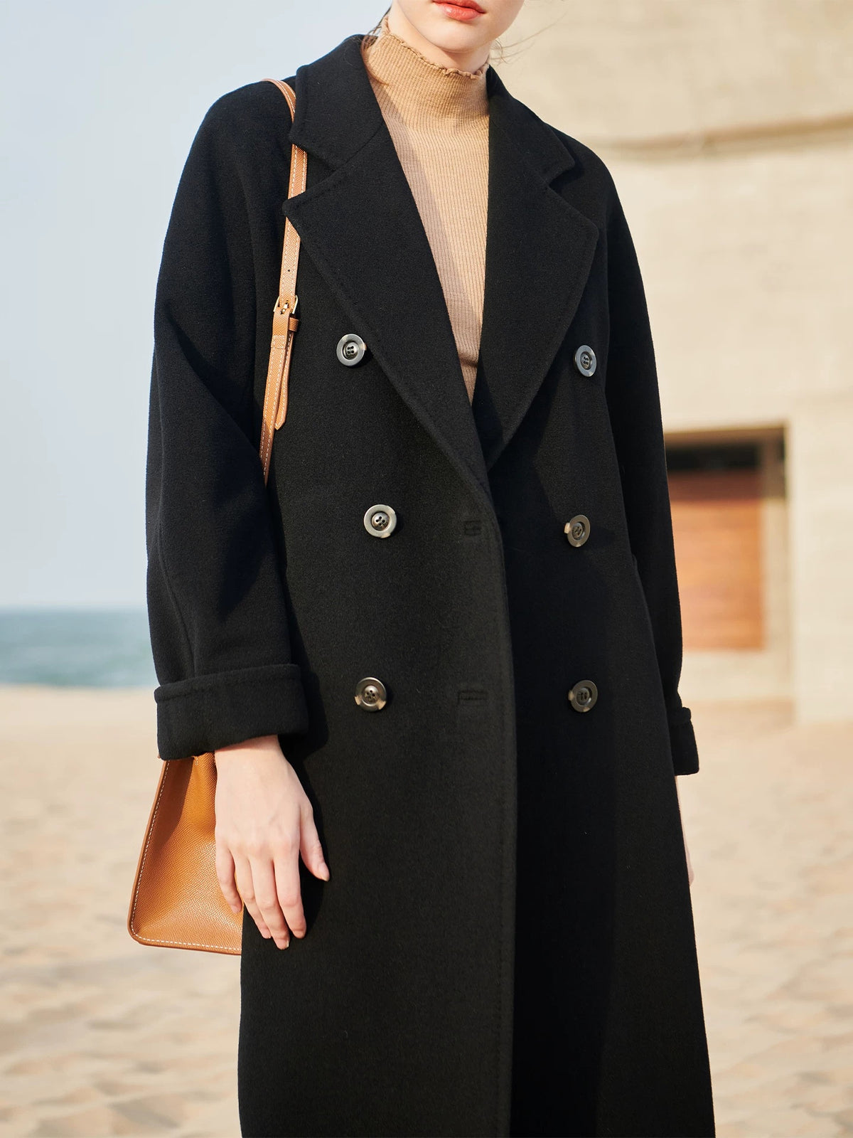 A woman wearing a long black double-breasted coat with silver buttons, styled with a beige turtleneck and a tan shoulder bag, standing outdoors near a sandy beach and stone building.
