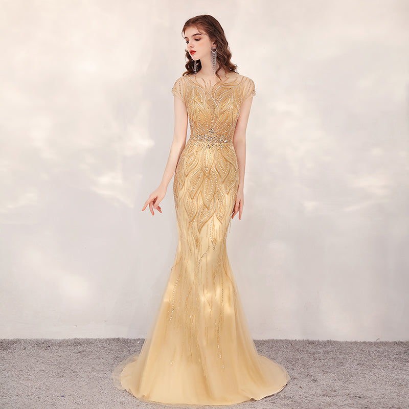 Woman wearing a gold evening gown with intricate detailing on a neutral background