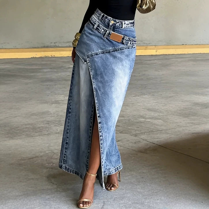 A model wearing a high-waisted, light blue denim maxi skirt with a high side slit, styled with a black top and gold heeled sandals in a concrete setting.