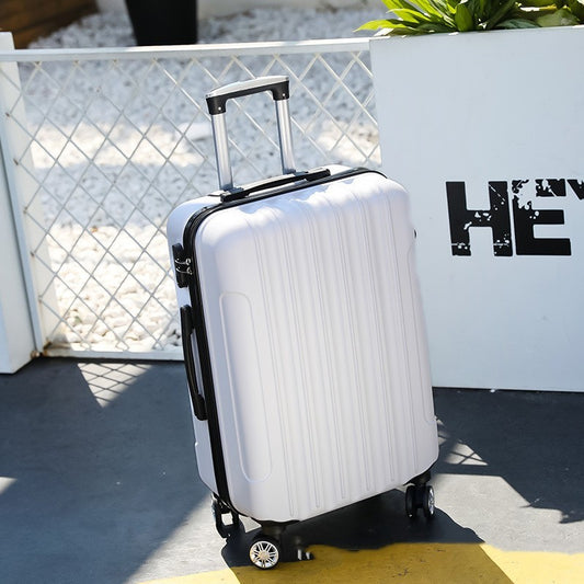 White suitcase with black handle and wheels on a concrete surface with a plant and white container in the background.