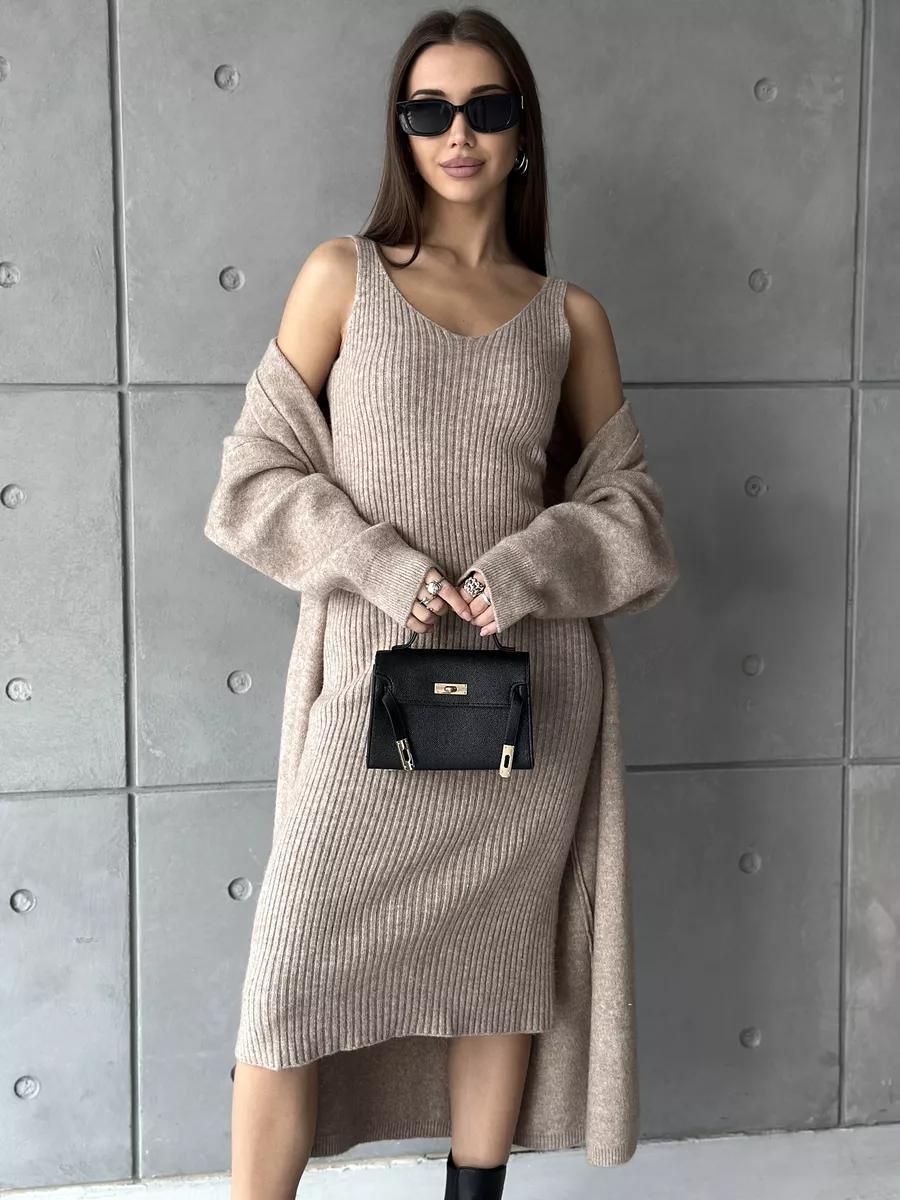 Woman wearing a long beige sweater dress against a gray tiled wall.